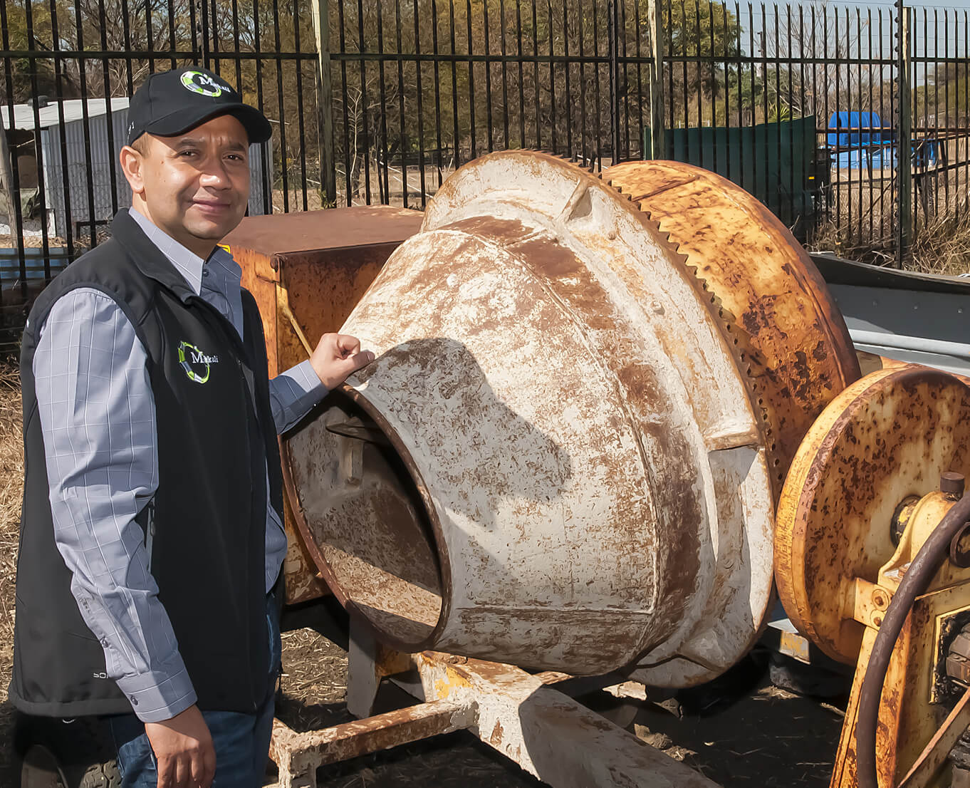 A man stands beside a large cement mixer, overseeing the construction process in a busy work environment.